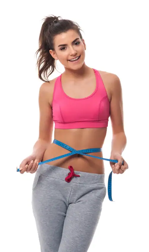 A woman in a pink sports bra and gray sweatpants measuring her waist with a blue tape measure.