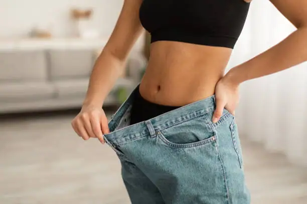 Slim woman in black sports bra and oversized jeans showing weight loss progress.