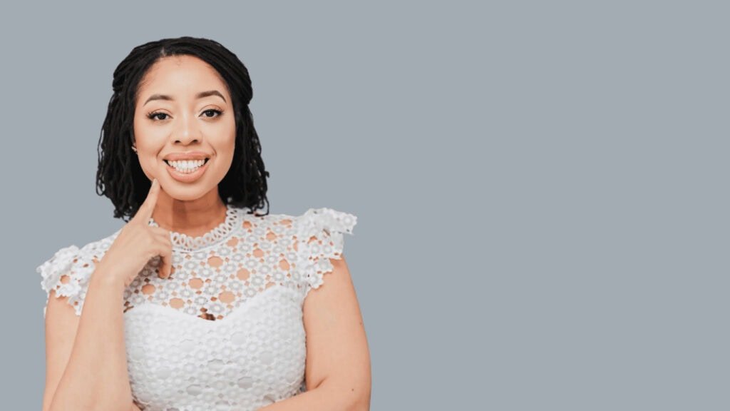 Woman smiling with finger on cheek, wearing white lace top, against light gray background.