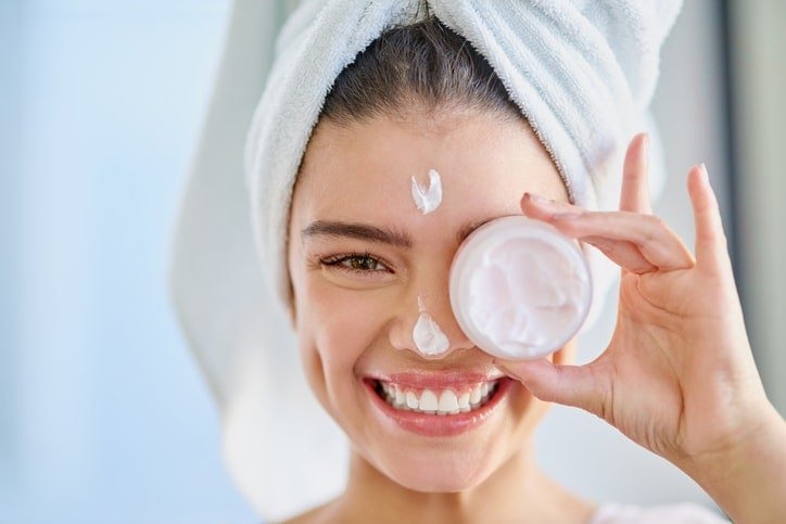 Clear skin care routine with a smiling woman applying moisturizer to her face.