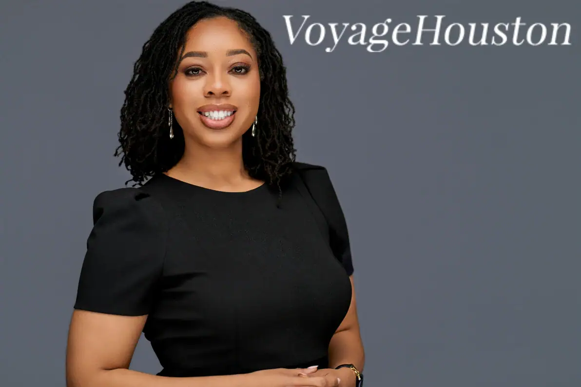 A woman with shoulder-length curly hair wearing a black top, smiling, with "VoyageHouston" text in t.