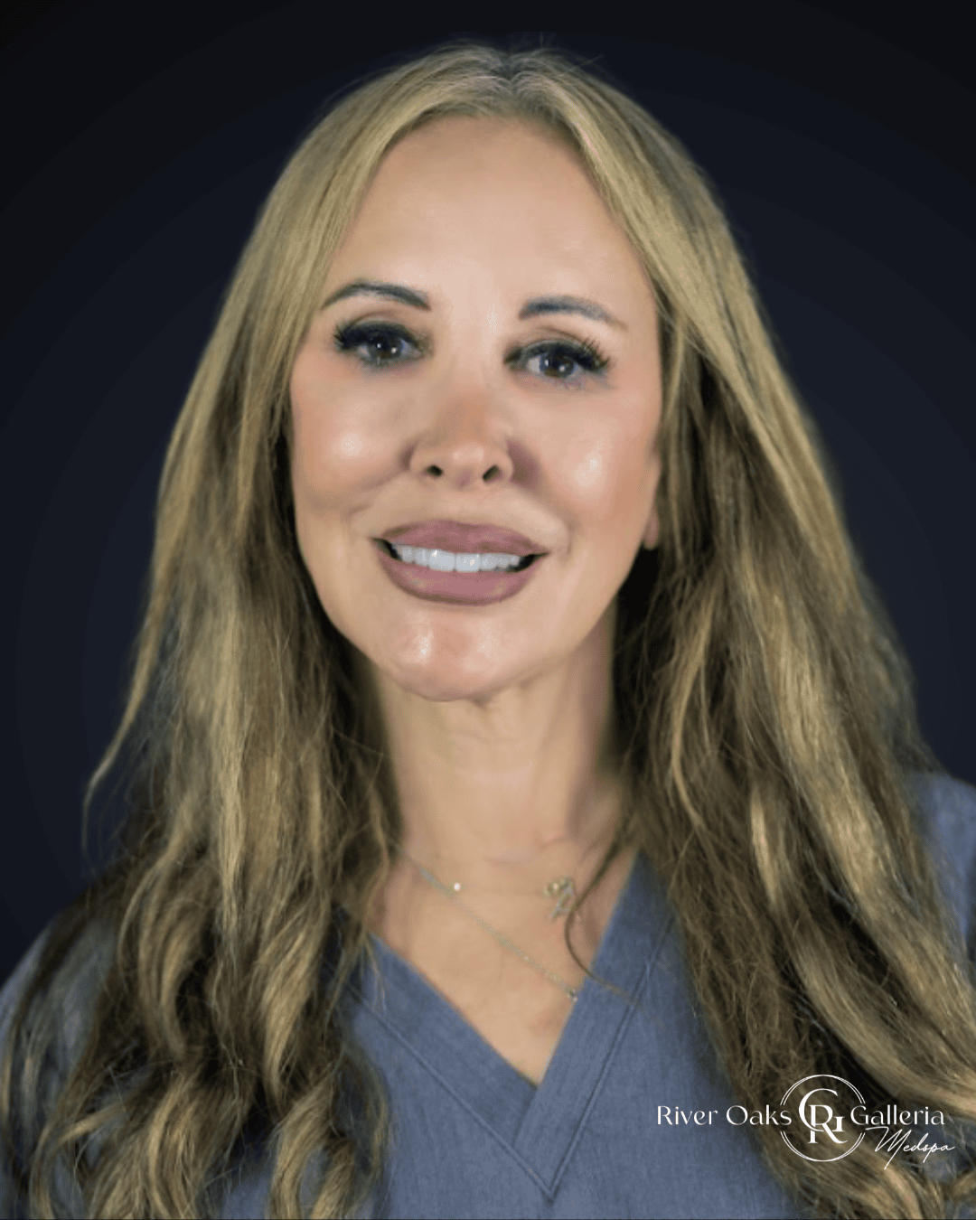 Woman with long blonde hair wearing blue scrubs, smiling, against dark background.